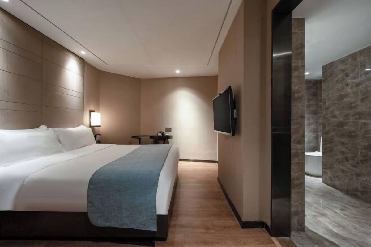 A modern hotel room with a large bed, a flat-screen TV mounted on the wall, a small table, and a nightstand.