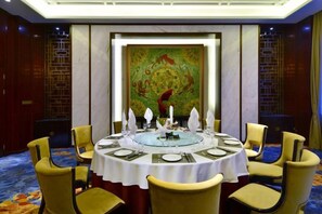 Une table de salle à manger ronde, recouverte d’une nappe blanche, entourée de chaises, dans une pièce où un grand tableau décoratif orne le mur.