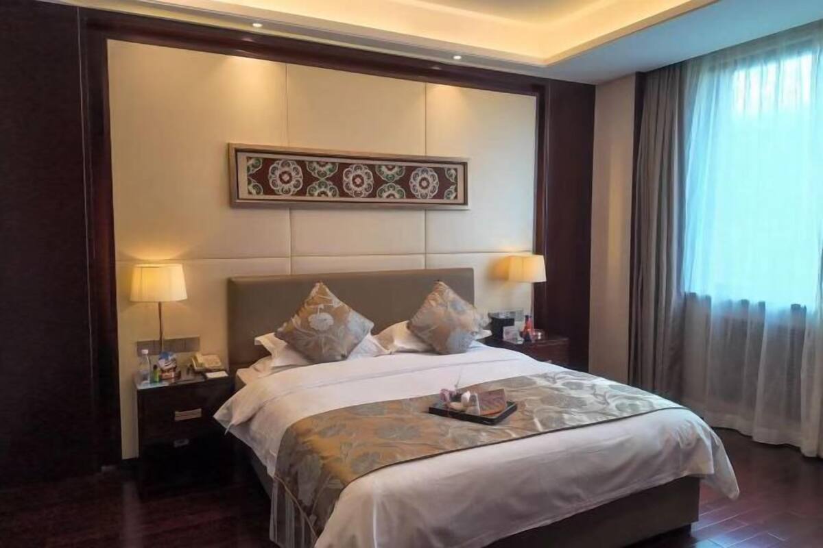 A hotel room with a large bed, two bedside lamps, a nightstand, a decorative wall panel, and a window with curtains.