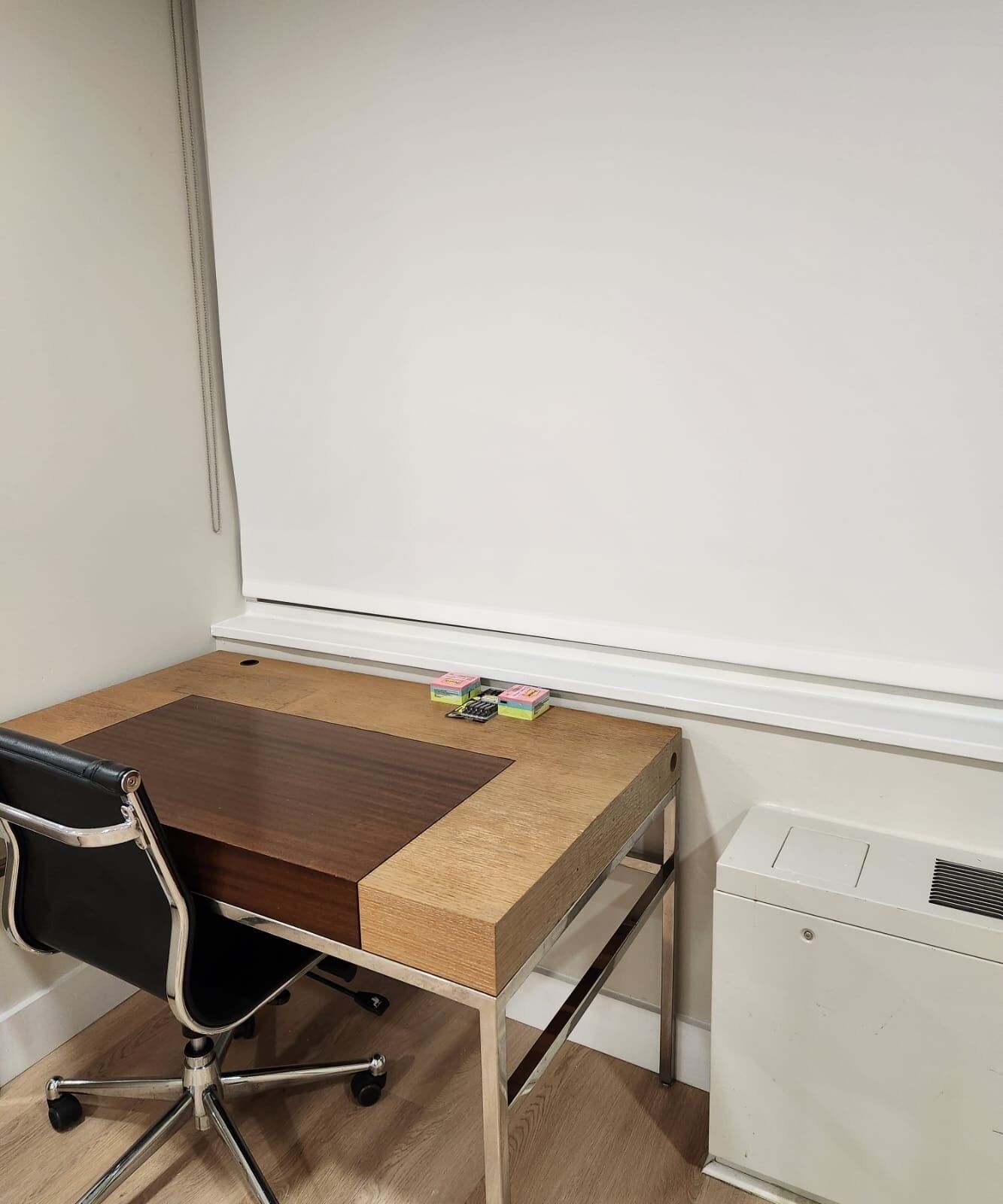 Um pequeno escritório com uma mesa de madeira, uma cadeira de escritório preta e um radiador branco.