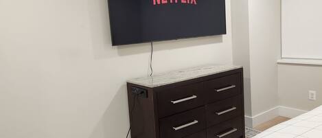 A bedroom with a flat-screen TV mounted on the wall displaying the Netflix logo, a dark wooden dresser with drawers, a patterned rug, and a bed with a white quilt.