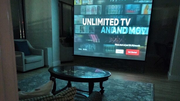 Ein Wohnzimmer mit einem großen Bildschirm, der eine Werbung für einen Fernsehservice zeigt. Im Raum befinden sich ein Sofa und ein Couchtisch.
