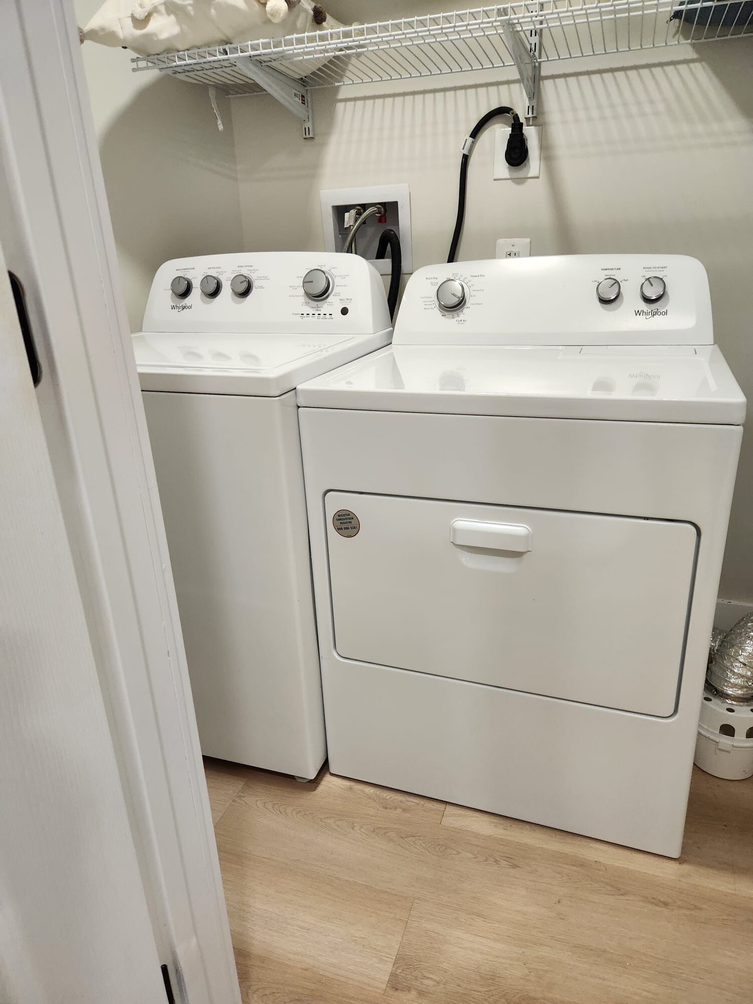 A laundry room with two white Whirlpool washers and dryers, a white shelf above, and a white door to the left.