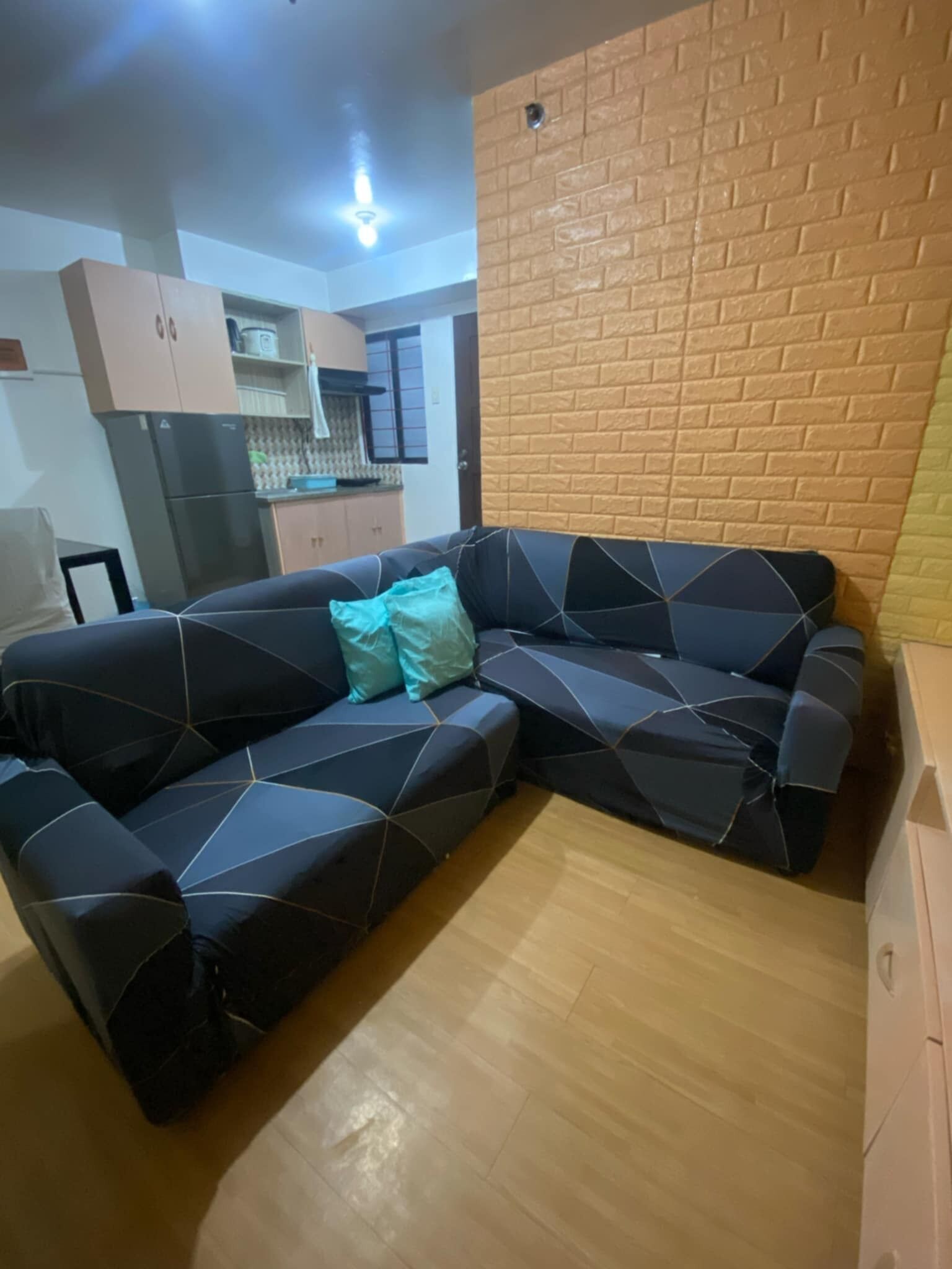 A living room with a sectional sofa, a kitchen area in the background, and a brick-patterned wall.