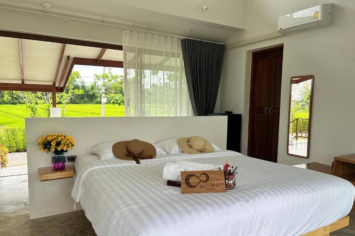 A bedroom with a large bed, a wooden headboard, a bedside table with a vase of flowers, and a view of a green landscape through a window.