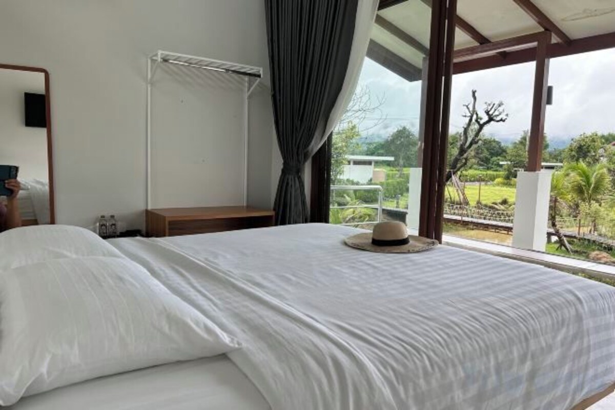 Une chambre à coucher avec un lit, une table de chevet, une fenêtre avec des rideaux et une vue sur la verdure à l’extérieur.