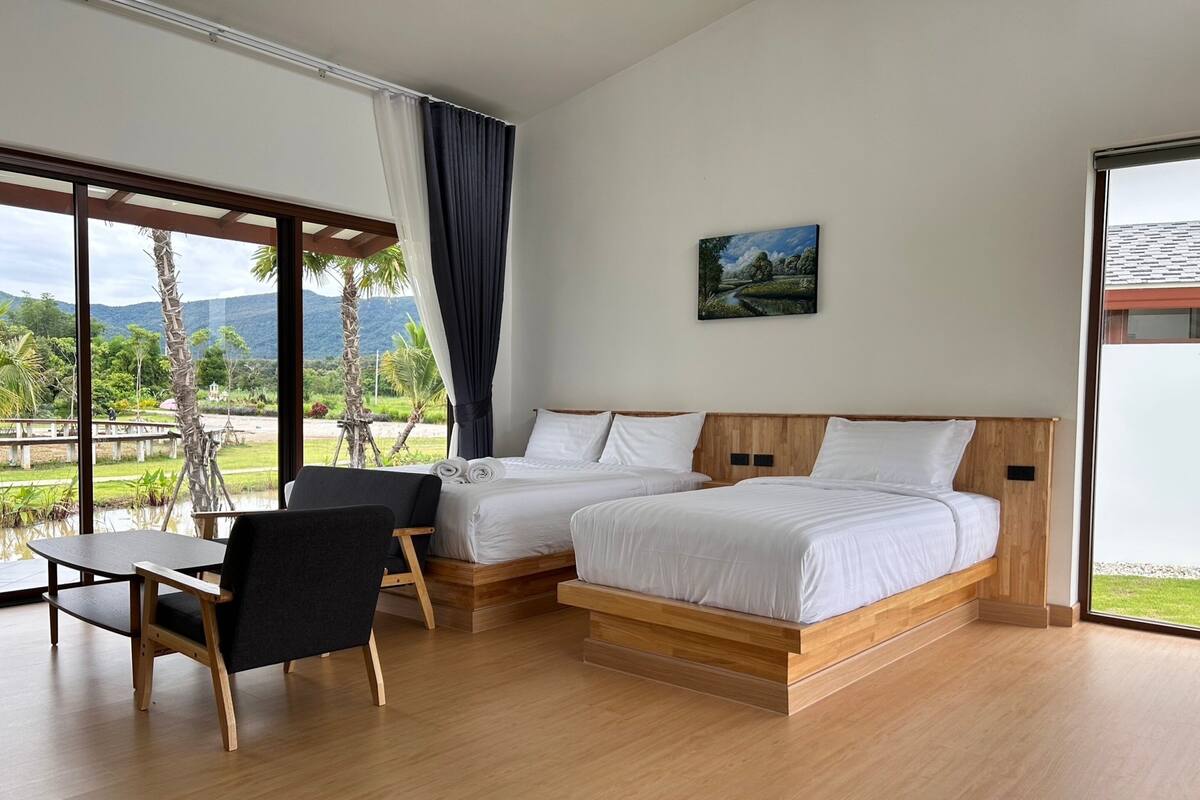 A modern bedroom with two beds, a desk, and a chair, featuring a large window with a view of greenery and mountains.