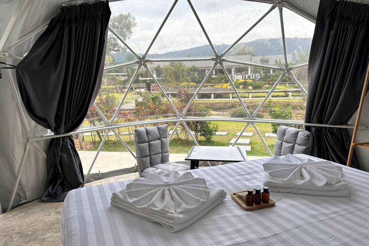 A room with a large bed, two chairs, a small table, and a view of a garden and mountains through a geometric window structure.