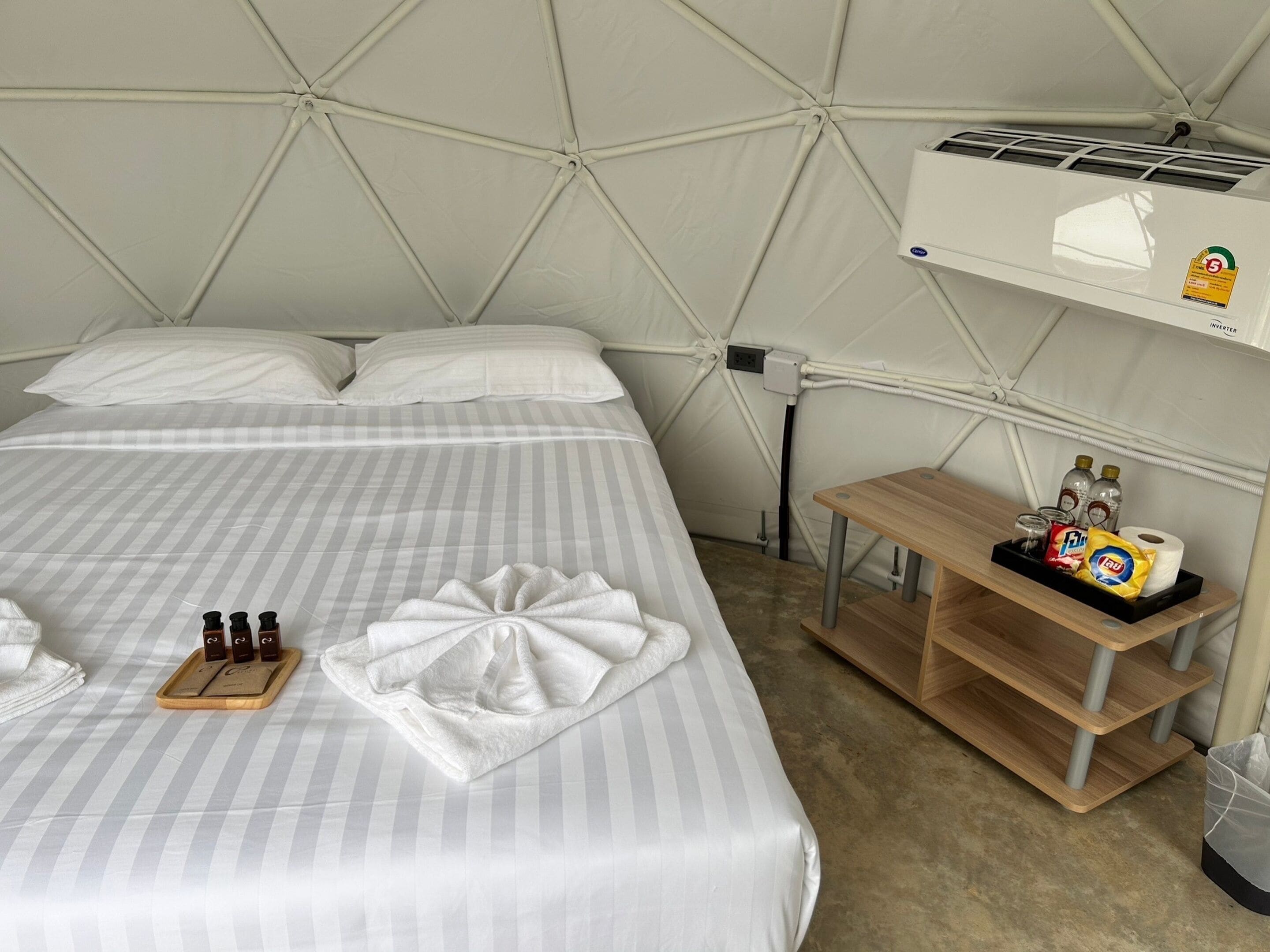 A tent-style room with a bed, pillows, a tray with toiletries, and a shelf with bottles and a roll of toilet paper.