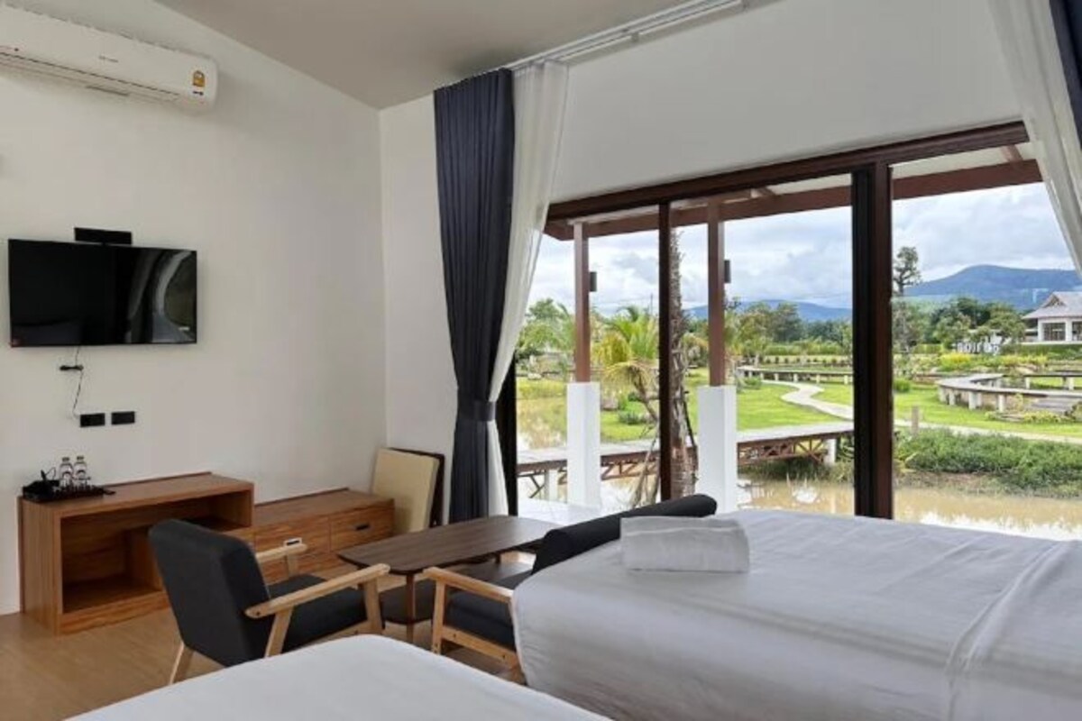 Une chambre d’hôtel équipée d’un lit, d’un bureau, d’une chaise, d’une télévision et d’une grande fenêtre donnant sur un jardin et des montagnes.