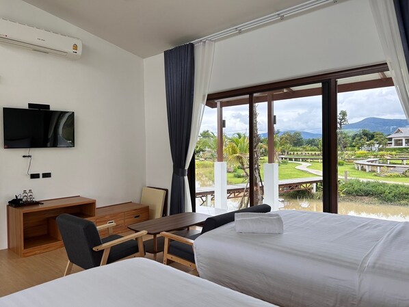 A hotel room with a bed, a desk, a chair, a TV, and a large window with a view of a golf course and mountains. - Sumator Resort (Chiang Rai)