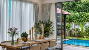 Un coin repas avec une table dressée pour quatre personnes, entouré de grandes fenêtres ornées de rideaux légers, offrant une vue sur une piscine et sur une végétation luxuriante à l’extérieur.