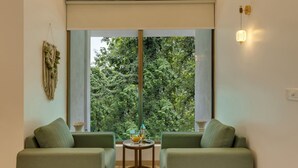 Une pièce avec deux canapés verts, une petite table ronde avec des boissons, une grande fenêtre donnant sur des arbres et un luminaire fixé au mur.