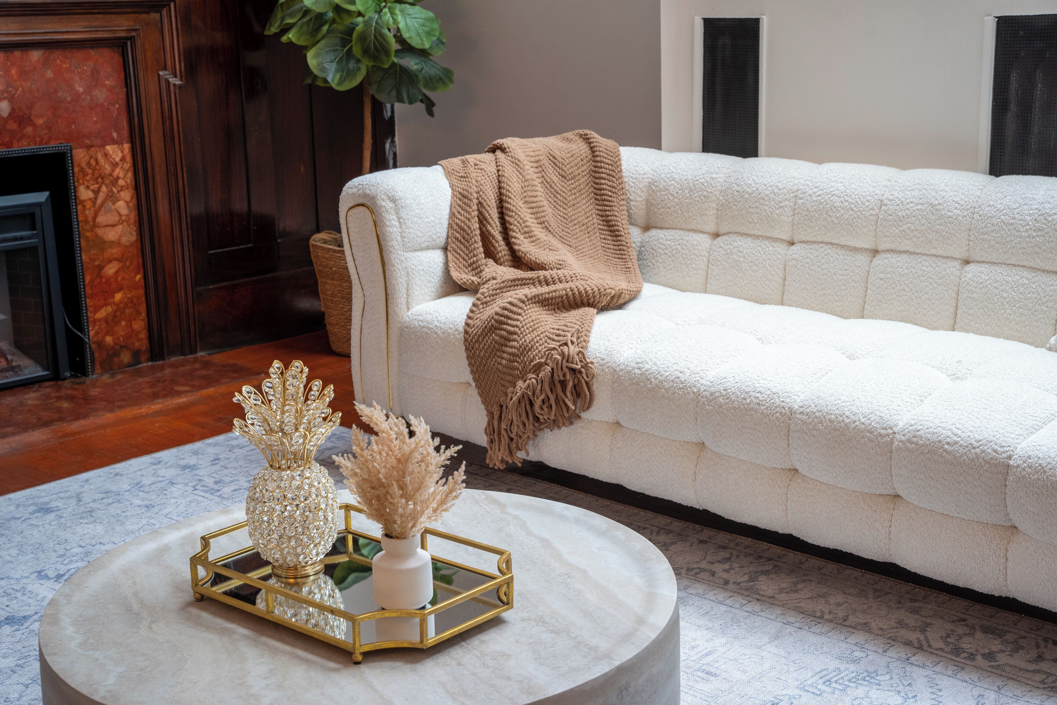 Un sofá blanco con tela texturizada, una mesa redonda de centro con una bandeja dorada y objetos decorativos, y una planta verde grande al fondo.