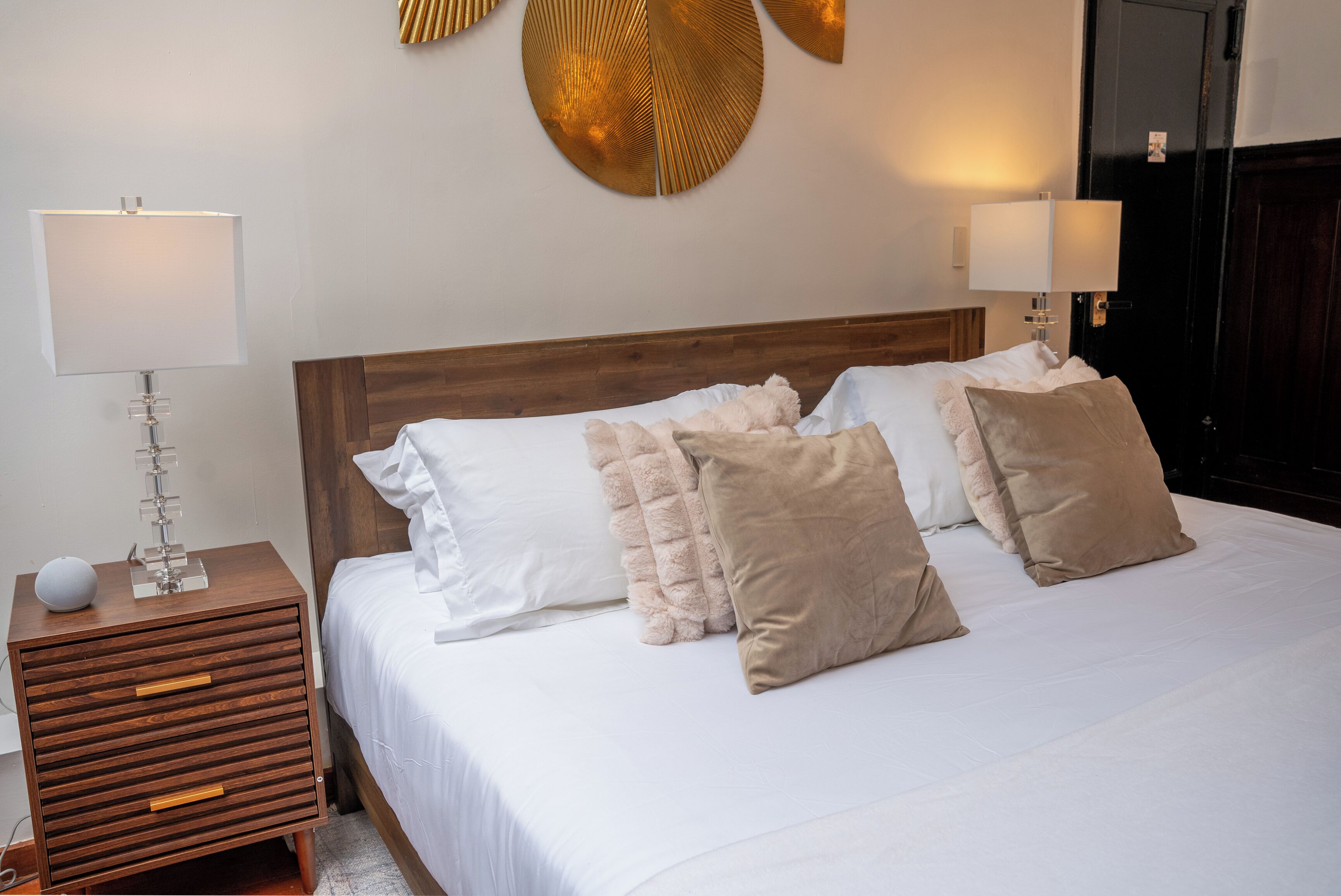 Una cama bien tendida con almohadas blancas y beige, una mesita de noche de madera con una lámpara, y una pared con un adorno circular.
