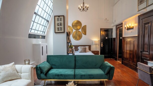 A spacious living area with a green sofa, a white armchair, and a round coffee table. A ladder is propped against the wall, and a bed is visible in the background. - Luxurious Loft Suite at Mansfield, Central NYC Location near Top Attraction (New York)