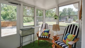 A sunroom with large windows, a wooden chair with colorful cushions, a small table, and a birdhouse decoration.