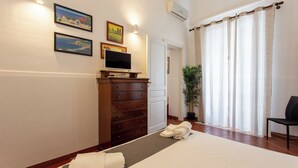 A hotel room with a bed, a TV on a dresser, a potted plant, and a view of the outdoors through the window. - Le dimore del mito - Atalanta (Syracuse)