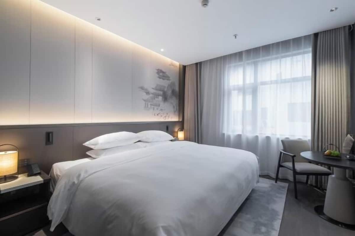 A modern hotel room with a large bed, a desk with a chair, and a view of the outdoors through large windows.