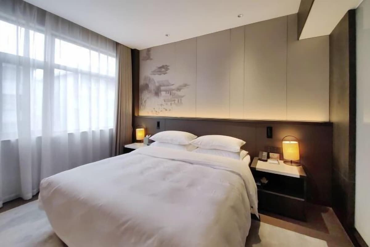 A hotel room with a large bed, two bedside tables with lamps, a wall-mounted artwork, and a window with sheer curtains.