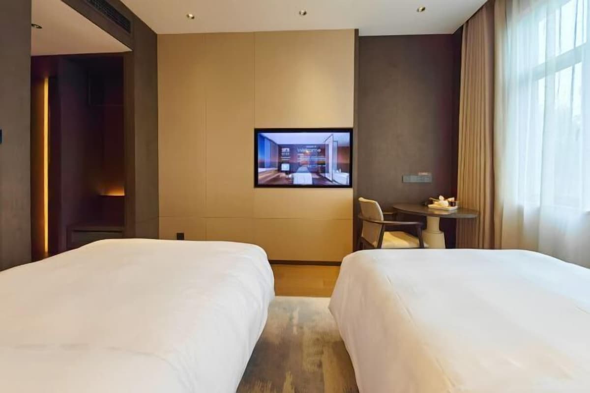 A hotel room with two beds, a television mounted on the wall, a small table with a chair, and a window with curtains.