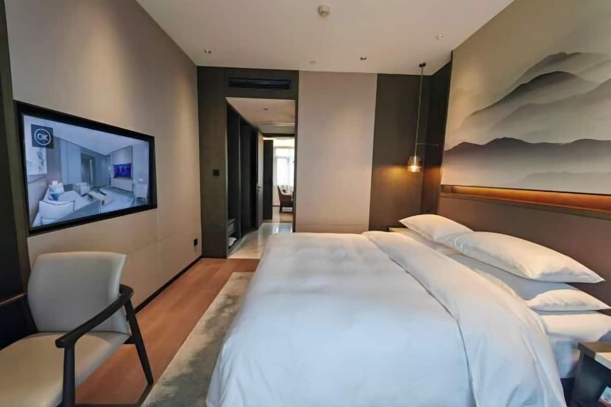 A modern hotel room with a large bed, a television mounted on the wall, and a view of the hallway.