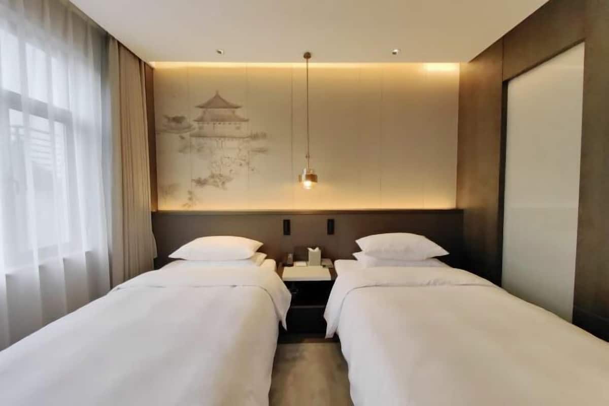 A hotel room with two single beds, a nightstand, a wall-mounted lamp, and a large window with sheer curtains.