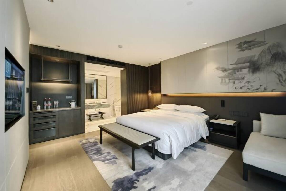 A modern bedroom with a large bed, bedside tables, a bench, a desk, and a wall-mounted TV.