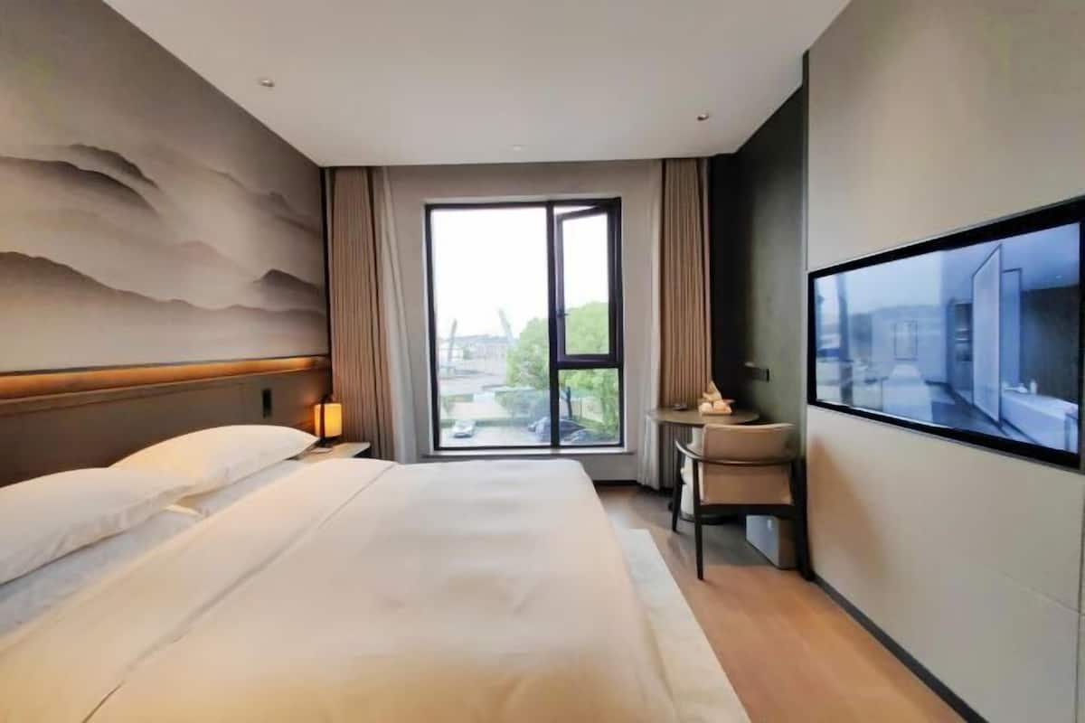 A hotel room with a large bed, a TV, a desk, and a view of the city through a large window.