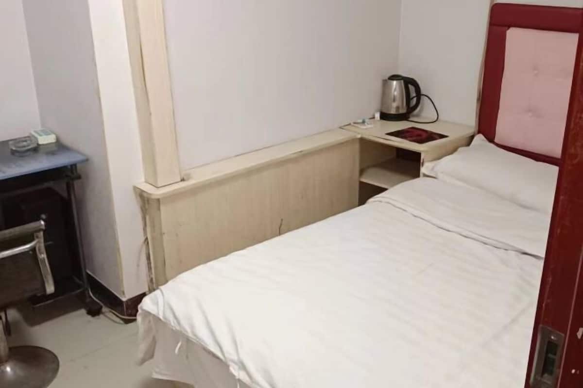 Una cama individual con colchón blanco y cabecera roja, un escritorio pequeño con teléfono, y una tetera sobre una mesita.