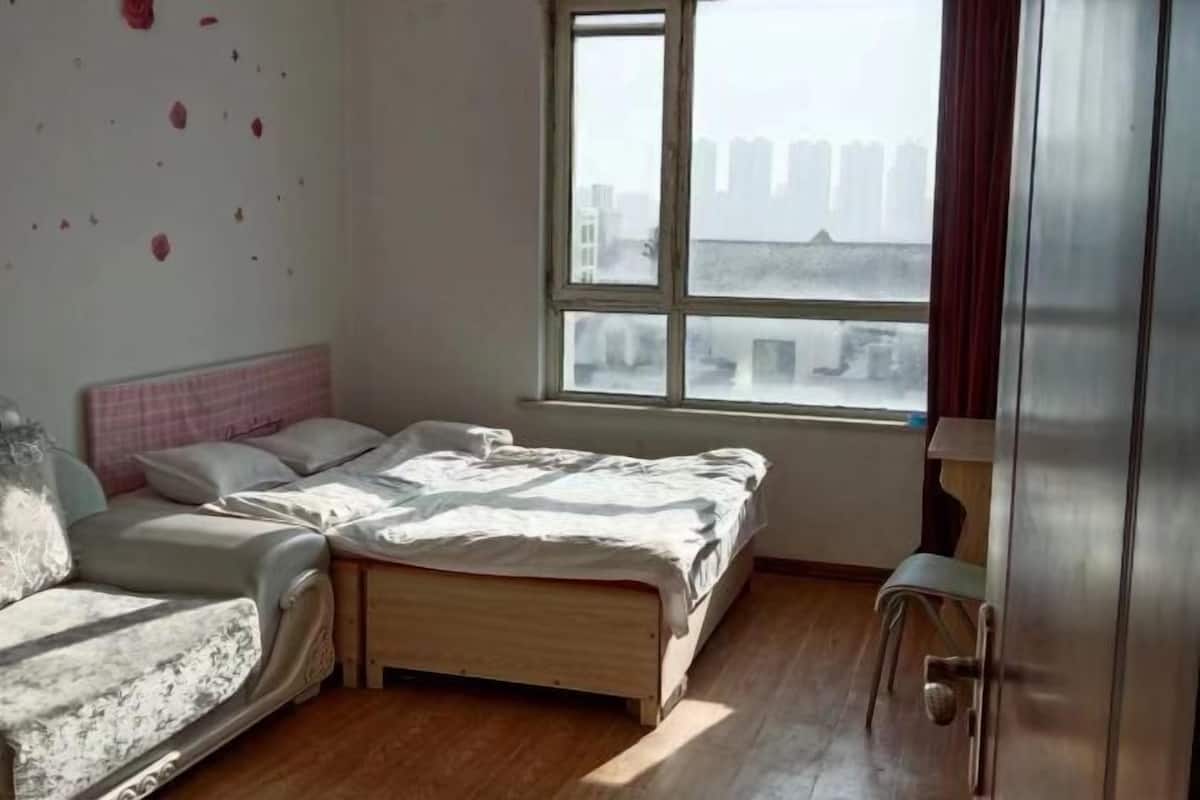 Un dormitorio con cama, sofá, ventana con vistas a edificios y una puerta.