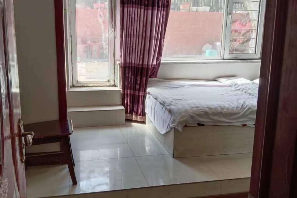 Un dormitorio con cama, ventana con cortinas, un taburete de madera y vistas al exterior.
