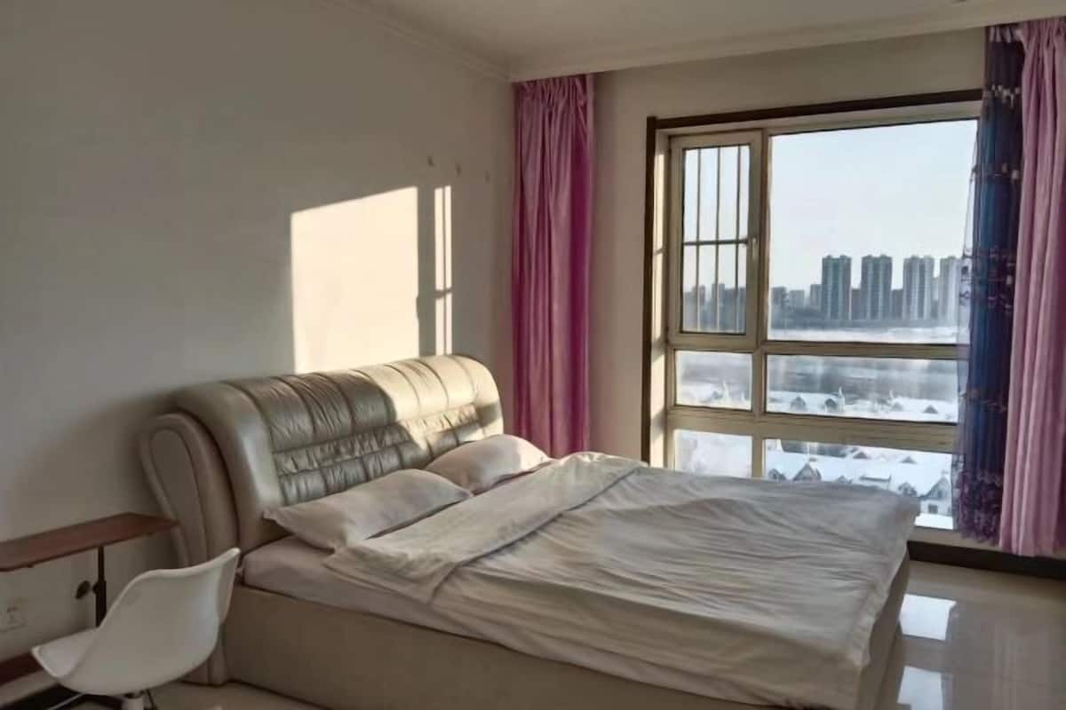 Un dormitorio con cama, una silla, una ventana con cortinas y vistas a edificios.