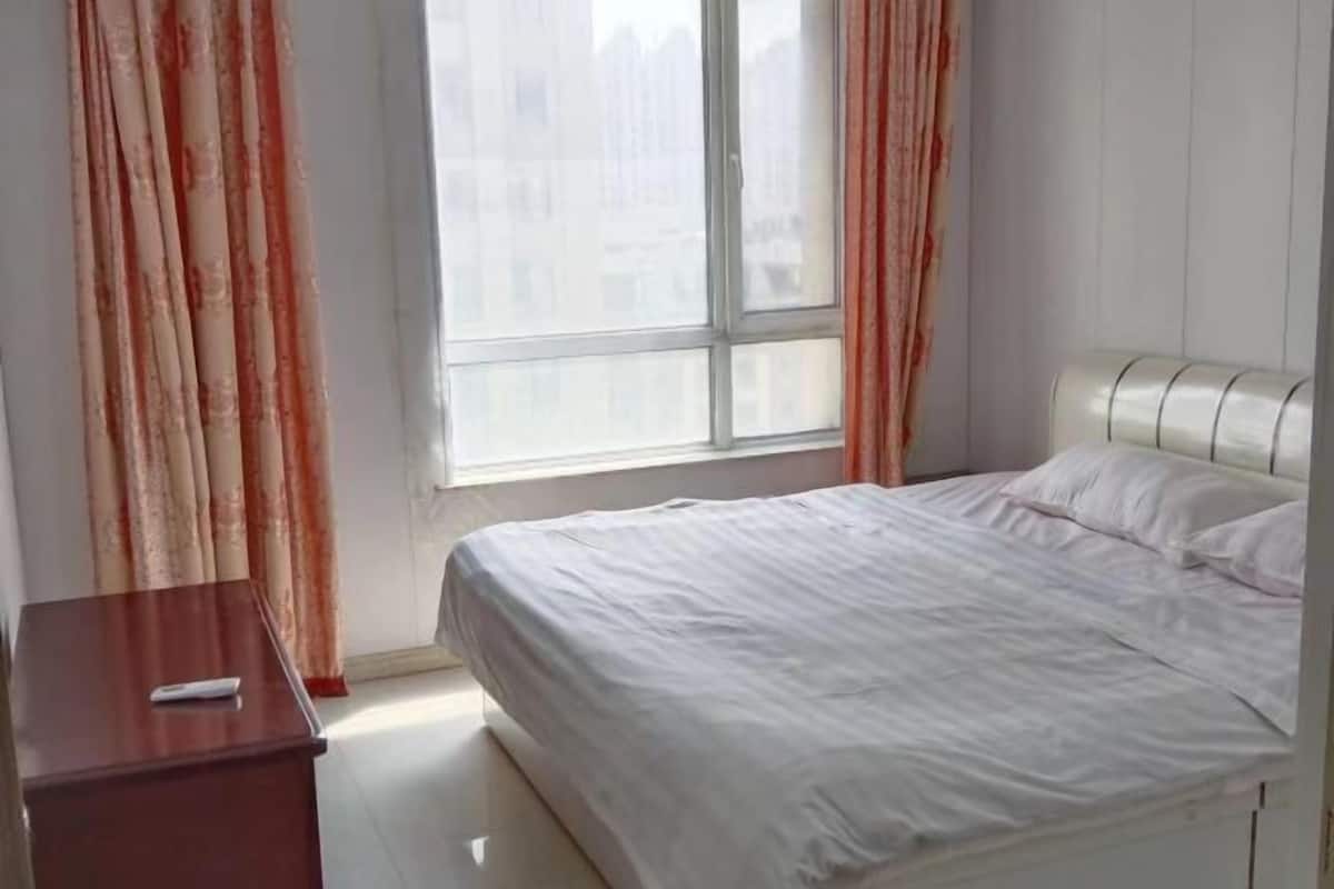Un dormitorio con cama, una mesita de noche, una ventana con cortinas y vistas a edificios.