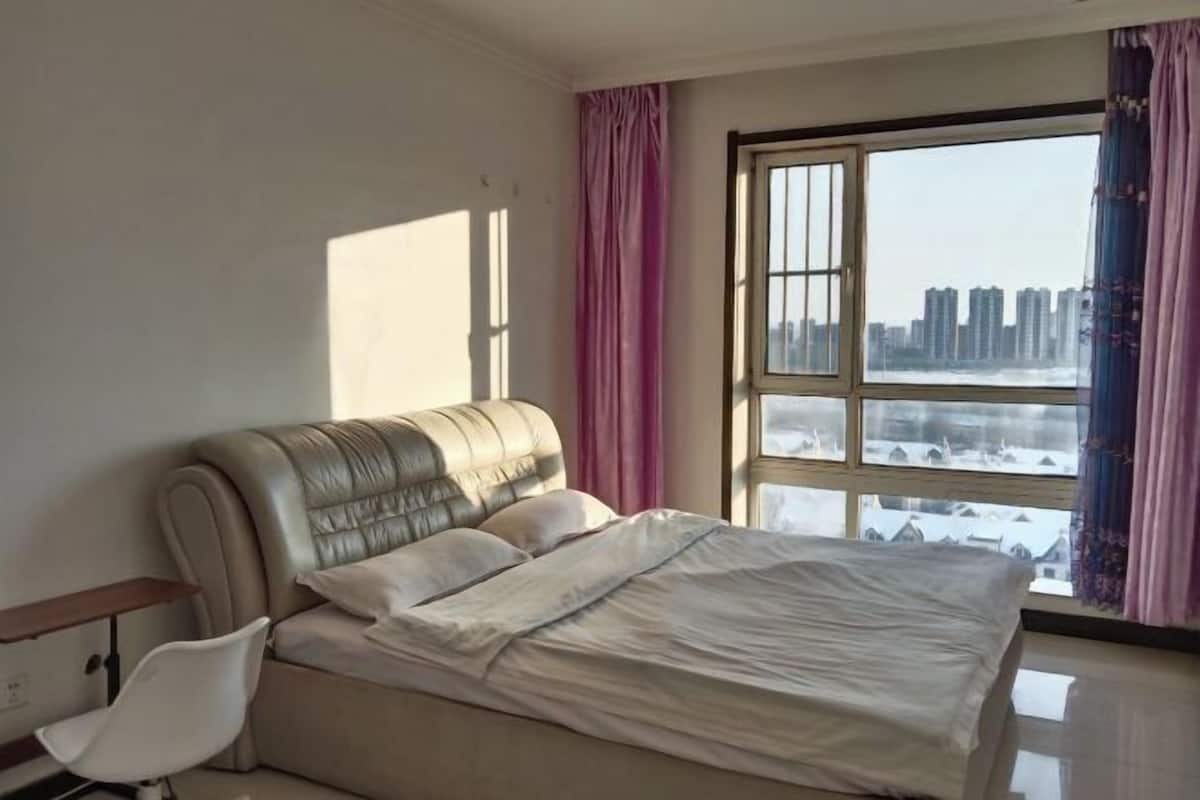 Un dormitorio con cama, una silla, un ventanal con cortinas y vistas a edificios.