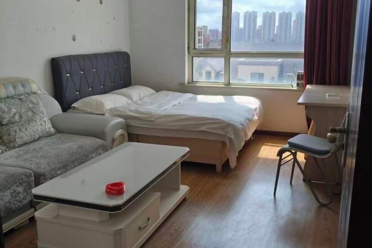A bedroom with a bed, a sofa, a small table, and a window with a view of buildings.