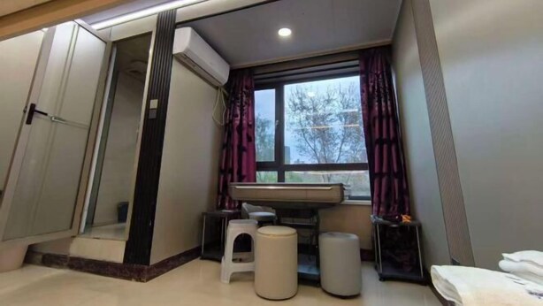 Habitación de hotel con cama, un taburete pequeño, ventana con cortinas, equipo de aire acondicionado y un espejo.
