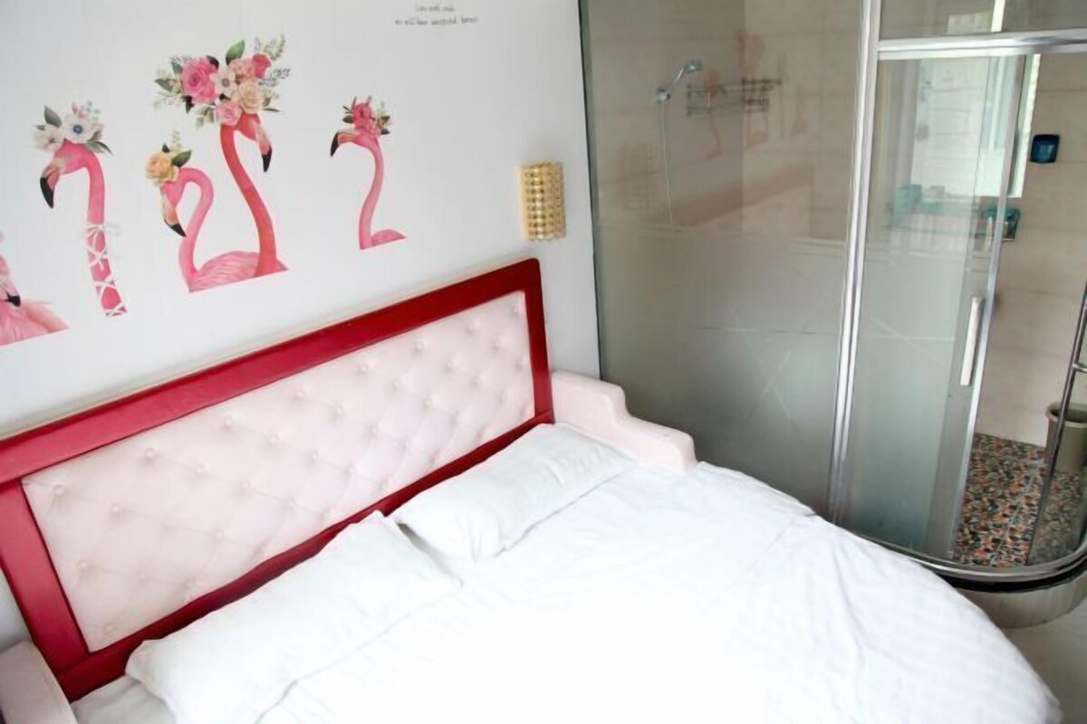 A bedroom with a red headboard, white bedding, a glass shower enclosure, and a wall with floral artwork.