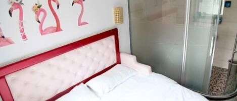 Une chambre à coucher avec une tête de lit rouge, du linge de lit blanc, une cabine de douche en verre et un mur orné d’œuvres d’art florales.
