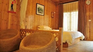 Une pièce de style cabane en bois, avec un lit, deux fauteuils en osier, un tableau accroché au mur et une fenêtre avec des rideaux.