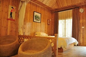 Une pièce de style cabane en bois, avec un lit, deux fauteuils en osier, un tableau accroché au mur et une fenêtre avec des rideaux.