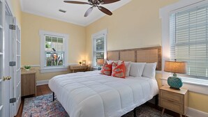 A bedroom with a large bed, a ceiling fan, a nightstand with a lamp, and a window with blinds. - Casa Amarillo l Steps to Historic District (St. Augustine)