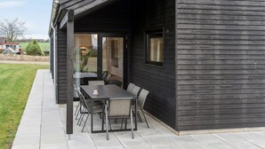 Um pátio externo com mesa e cadeiras, uma porta de vidro que dá para um jardim, e fachada de madeira escura.
