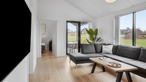 Uma sala de estar moderna com um sofá cinzento, uma mesa de centro de madeira e uma janela grande com vista para o exterior.