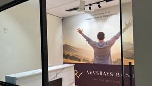 Et receptionsområde med et plakatbillede af en mand med armene i vejret, udsigt til bjerge, og teksten "Every Stay is a Celebration of Happiness" og "VoVSTAYS BANU THE NESTLED ONE".