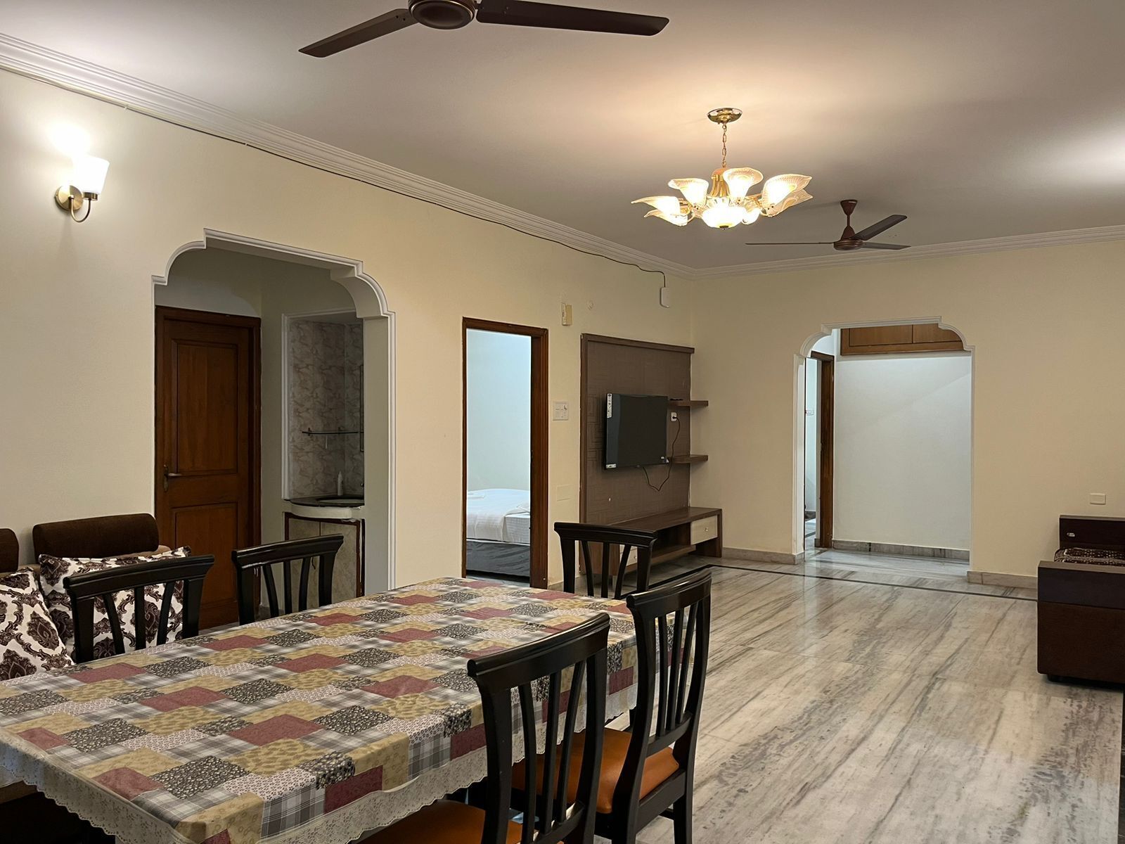 A dining area with a table set for four, a television mounted on the wall, and a ceiling fan.