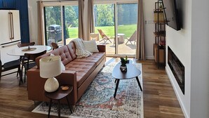 Uma sala de estar espaçosa com um sofá grande, uma mesa de centro e uma TV fixada na parede. Há uma porta de vidro deslizante que dá para um pátio com móveis externos.