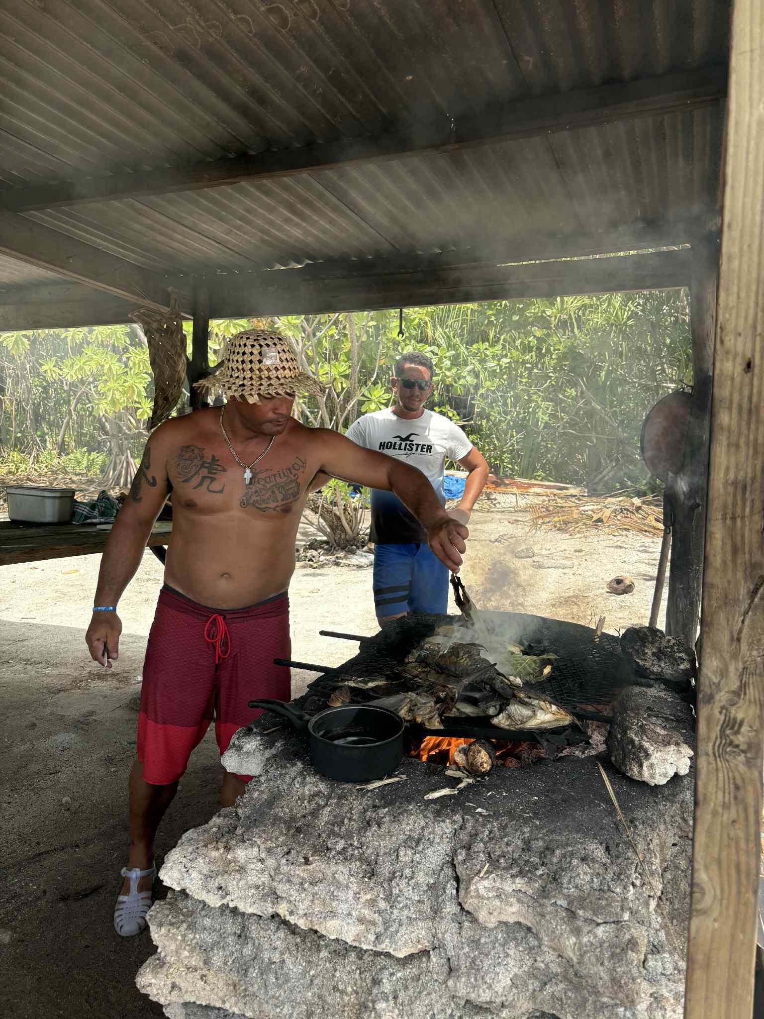 Une personne fait griller du poisson sur un feu de camp, à l’abri sous une structure en bois.