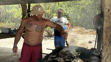 Une personne fait griller du poisson sur un feu de camp, à l’abri sous une structure en bois.