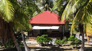 A building with a red roof surrounded by palm trees and small bushes. - Location Maison Entière a 5 Minutes à Pied D'une Plage (Rangiroa)